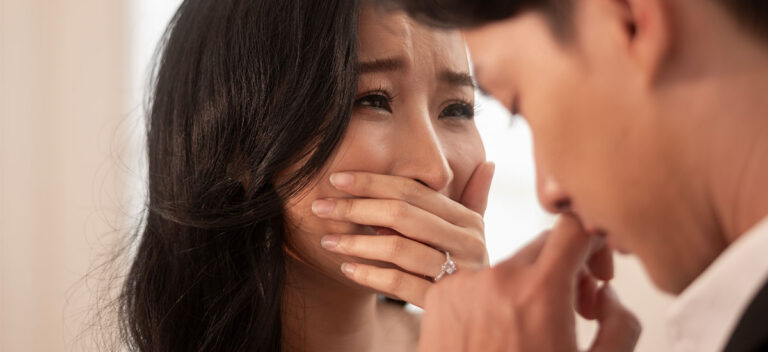 A woman with long dark hair holds a hand over her mouth with a tearful, distressed expression, sitting across from a man shown from behind in soft focus.